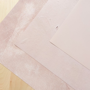 May include: Three pieces of textured paper in shades of pink, arranged in a diagonal pattern on a light wood surface.