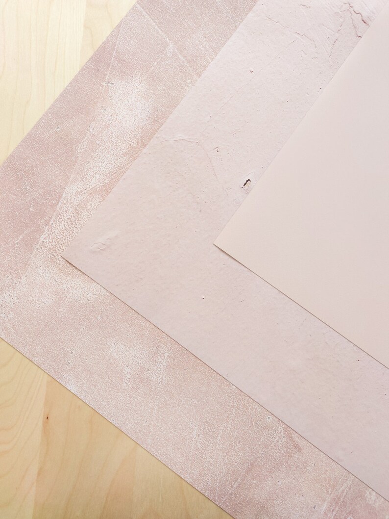 May include: Three pieces of textured paper in shades of pink, arranged in a diagonal pattern on a light wood surface.