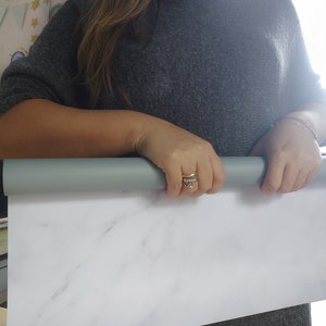 May include: A person holds a rolled up white marble-patterned backdrop. The backdrop is made of a smooth, white material with gray veining.