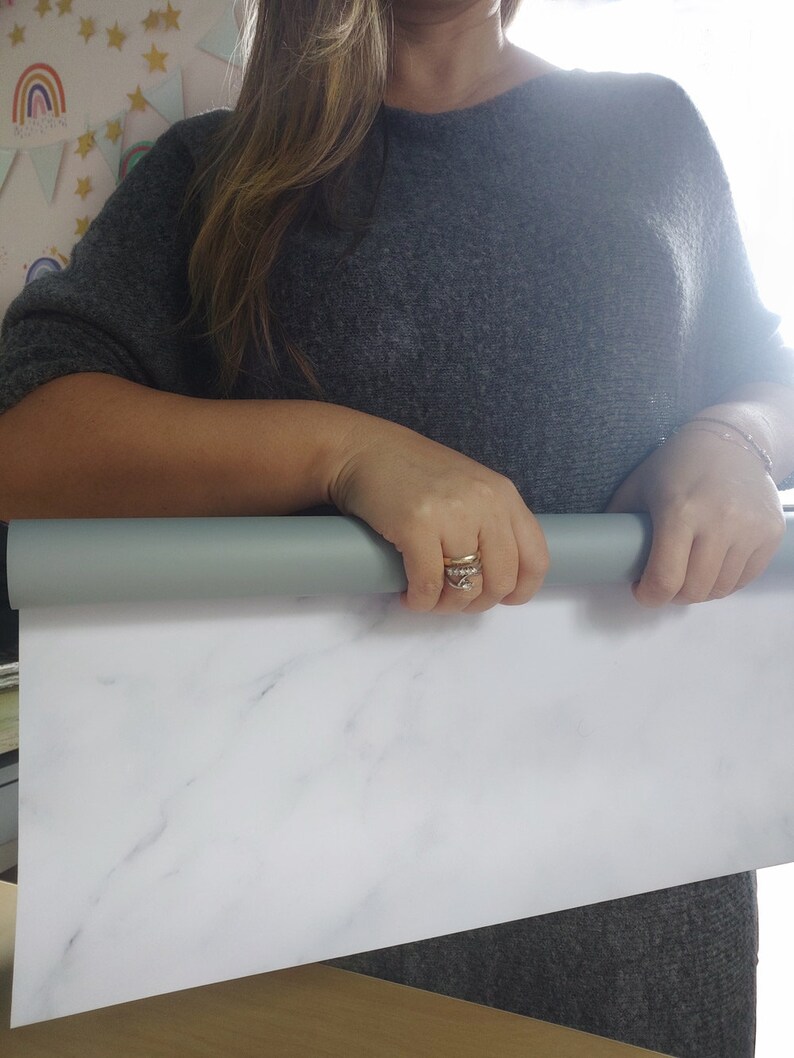 May include: A person holds a rolled up white marble-patterned backdrop. The backdrop is made of a smooth, white material with gray veining.