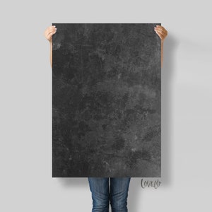 Peut inclure: Une grande toile rectangulaire gris foncé avec une surface texturée et mouchetée. La toile est tenue par une personne, et le bas du jean de la personne est visible. La toile a un aspect industriel vieilli.