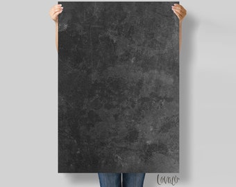 May include: A large, rectangular, dark grey canvas with a textured, mottled surface. The canvas is held up by a person, and the bottom of the person's jeans are visible. The canvas has a distressed, industrial look.