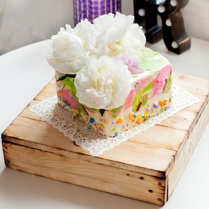 May include: A square cake decorated with colourful sprinkles and white peonies, sitting on a wooden cake stand.