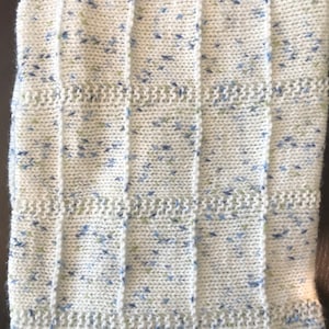 Op de afbeelding: Een wit gebreid babydekentje met blauwe en groene vlekjes. Het dekentje heeft een geribbelde textuur.