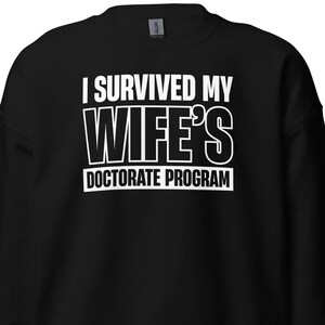 May include: Black sweatshirt with white text that reads "I survived my wife's doctorate program."