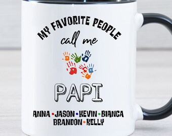 May include: White ceramic coffee mug with a black rim and handle. The mug has a black design that says "My favorite people call me Papi" with a circle of colorful handprints and the names "Anna, Jason, Kevin, Bianca, Brandon, Kelly" below.