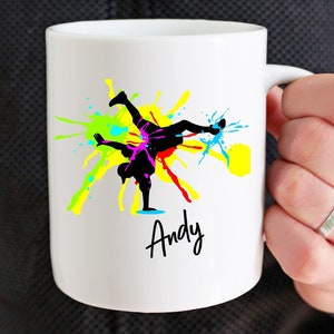 May include: White ceramic mug with a black silhouette of a person breakdancing with colorful paint splatters in the background. The name "Andy" is written in black cursive below the image.