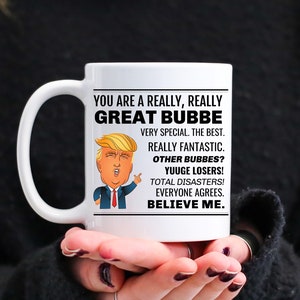 May include: White ceramic mug with a black handle. The mug features a cartoon image of a man with blonde hair and a red tie. The text on the mug reads "You are a really, really Great Bubbe. Very special. The best. Really fantastic. Other Bubbes? Yuuge Losers! Total Disasters! Everyone agrees. Believe me."