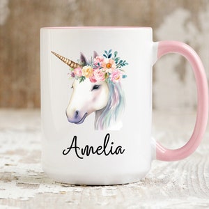 May include: White ceramic mug with a pink rim and handle. The mug features a watercolor illustration of a unicorn with a floral crown and the name "Amelia" printed below.