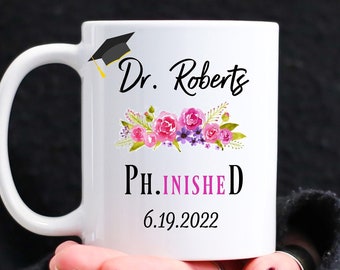 May include: White ceramic mug with a black graduation cap, pink flowers, and the text "Dr. Roberts PH.INISHED 6.19.2022".