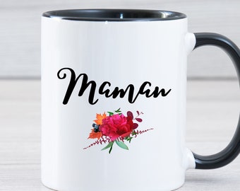 May include: White ceramic mug with a black rim and handle. The mug has a floral design and the word "Maman" in black script.