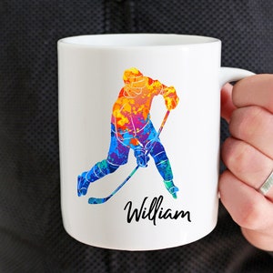 May include: White ceramic coffee mug with a colorful watercolor illustration of a hockey player and the name "William" printed on the bottom.