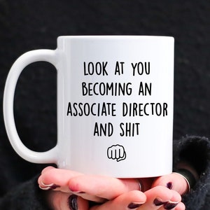 May include: White ceramic mug with black text that reads "LOOK AT YOU BECOMING AN ASSOCIATE DIRECTOR AND SHIT" with a fist graphic. The mug has a large handle and is held by a person.