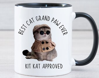 May include: White ceramic mug with a black rim and handle. The mug features a cartoon cat wearing sunglasses and a hat with the text "Best Cat Grand Paw Ever" and "Kit Kat Approved" printed on the mug.