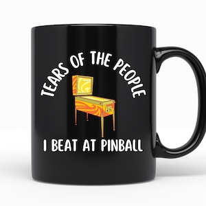 May include: Black ceramic mug with white text that reads "Tears of the people I beat at pinball". A cartoon image of a pinball machine is in the center of the mug.
