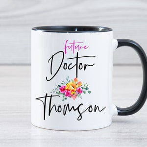 May include: White ceramic mug with a black rim and handle. The mug has a floral design and the text "future Doctor Thompson" in black and pink lettering.