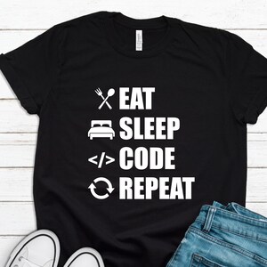 May include: Black t-shirt with the words "EAT SLEEP CODE REPEAT" in white, along with related icons. The shirt is paired with blue jeans and white sneakers, set against a white wood background.
