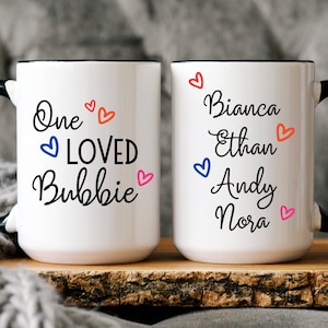 May include: Two white ceramic mugs with black handles. One mug says "One Loved Bubbie" with colorful hearts. The other mug says "Bianca Ethan Andy Nora" with colorful hearts.