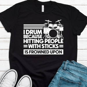 May include: Black t-shirt with white text and a drum set graphic. The text reads: "I DRUM BECAUSE HITTING PEOPLE = WITH STICKS = IS FROWNED UPON." The shirt is paired with blue jeans and white sneakers.
