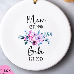 May include: A white ceramic Christmas ornament with a gold hanging loop. The ornament features the text "Mom EST. 1990" above a floral design in pink and purple hues, and the text "Bibi EST. 202X" below the flowers.