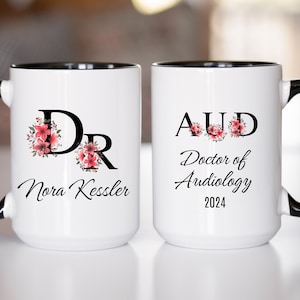 May include: Two white coffee mugs with black handles and black rims. The mug on the left has a floral design with the letters "DR" and the text "Nora Kessler". The mug on the right has a floral design with the letters "AUD" and the text "Doctor of Audiology 2024".
