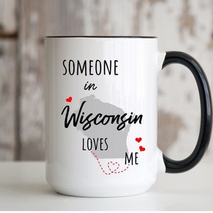 May include: White ceramic mug with a black rim and handle. The mug has a black graphic that says "Someone in Wisconsin loves me" with a red heart and a dashed line connecting the words "loves" and "me".