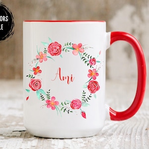 May include: White ceramic mug with a red handle and rim. The mug features a floral wreath design in pink and red, with the name "Ami" in the center. The words "OTHER COLORS AVAILABLE" are in a black circle.