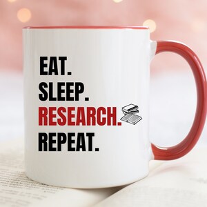 May include: White ceramic mug with a red rim and handle. The mug has black text that reads "Eat. Sleep. Research. Repeat." with a small illustration of a stack of books.