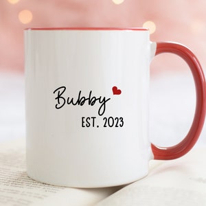 May include: White ceramic mug with a red rim and handle. The mug has black text that reads "Bubby EST. 2023" and a small red heart.