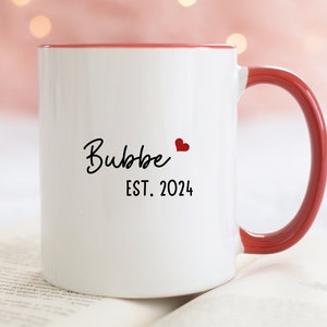 May include: White ceramic mug with a red rim and handle. The mug has black text that reads "Bubbe" with a small red heart and "EST. 2024" underneath.