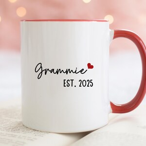 May include: White ceramic mug with a red rim and handle. The mug has black text that reads "Grammy EST. 2025" with a small red heart.