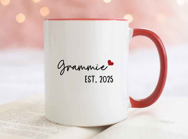 May include: White ceramic mug with a red rim and handle. The mug has black text that reads "Grammy EST. 2025" with a small red heart.