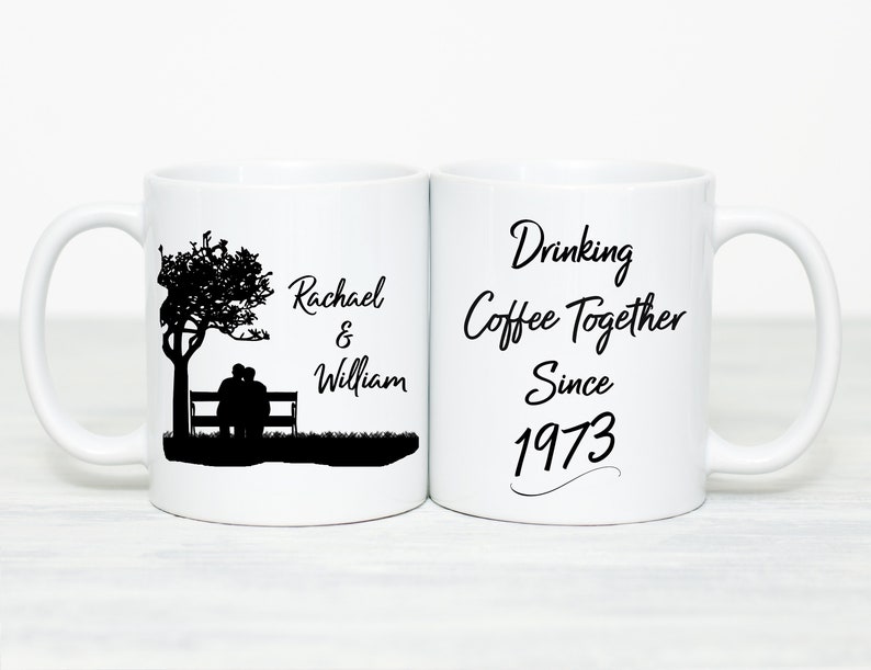 May include: Two white coffee mugs with black text and a silhouette of a couple sitting on a bench under a tree. One mug says "Rachael & William" and the other says "Drinking Coffee Together Since 1973".