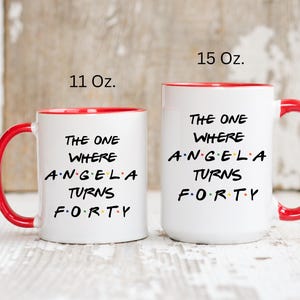 Op de afbeelding: Twee witte keramische mokken met rode binnenkant en handvatten. De mokken hebben de tekst "THE ONE WHERE ANGELA TURNS FORTY" in het zwart. E&eacute;n mok is 312 ml, de andere 425 ml.
