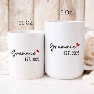 May include: Two white ceramic mugs with black text that reads "Grammy Est. 2025" and a small red heart. The mug on the left is 11 oz and the mug on the right is 15 oz.