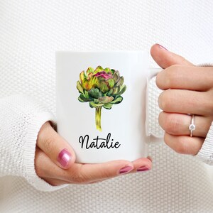 May include: White ceramic mug with a watercolor illustration of an artichoke and the name "Natalie" printed in black.