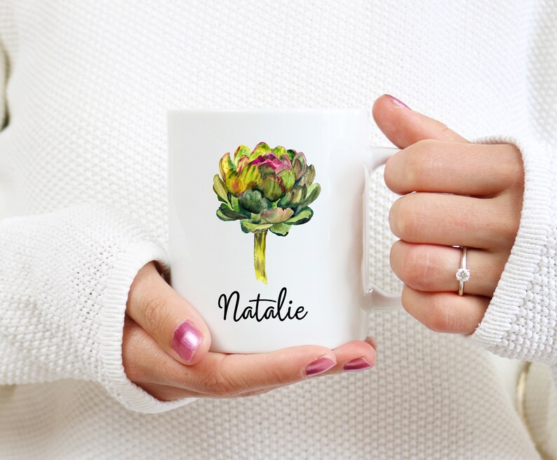 May include: White ceramic mug with a watercolor illustration of an artichoke and the name "Natalie" printed in black.