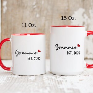 May include: Two white ceramic mugs with red rims and handles. The smaller mug is 11 ounces and the larger mug is 15 ounces. Both mugs have the text "Grammy Est. 2025" with a small red heart.