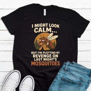 May include: Black t-shirt with the text "I MIGHT LOOK CALM... BUT I'M PLOTTING MY REVENGE ON LAST NIGHT'S MOSQUITOES." The shirt features a cartoon mosquito with a knife and fork. The shirt is paired with blue jeans and white sneakers.