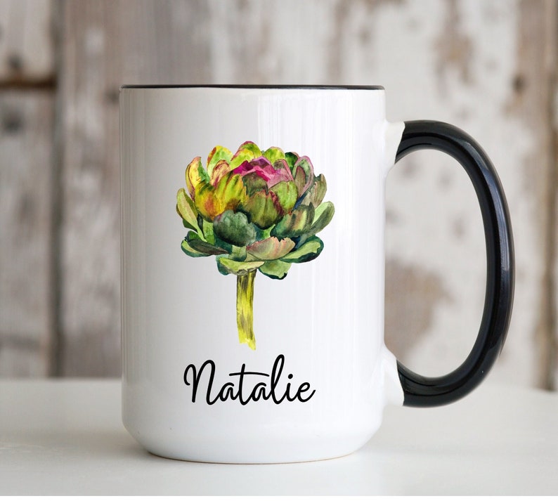 May include: White ceramic mug with a black handle. The mug features a watercolor illustration of an artichoke with green, yellow, and purple hues. The name "Natalie" is written in black cursive below the artichoke.