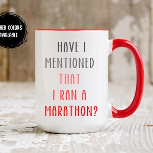 May include: White ceramic mug with a red interior and handle. The mug features the text "HAVE I MENTIONED THAT I RAN A MARATHON?" in gray and red lettering. A black circle with the text "OTHER COLORS AVAILABLE" is in the upper left corner.