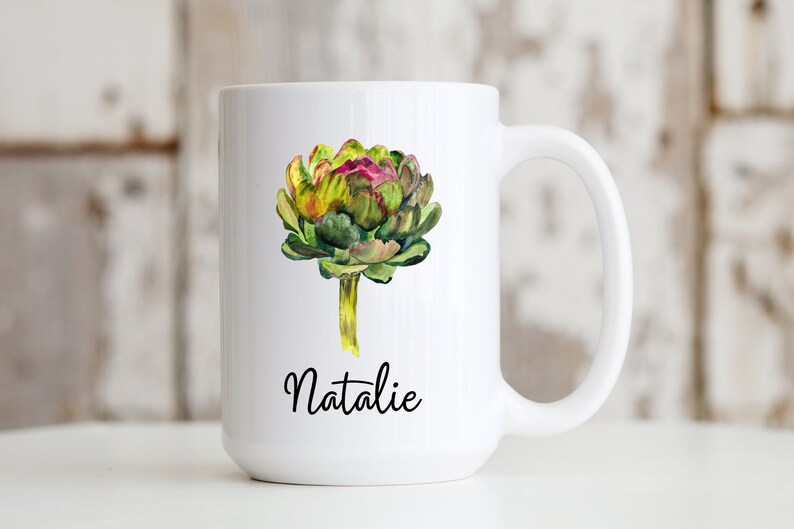 May include: White ceramic mug with a watercolor illustration of an artichoke in shades of green, yellow, and purple. The name "Natalie" is written in black cursive below the artichoke.