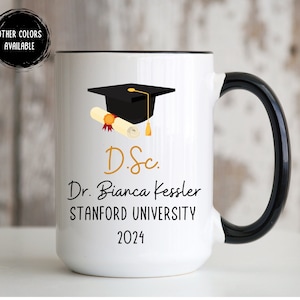 May include: White ceramic coffee mug with a black rim and handle. The mug features a black graduation cap with a gold tassel, a gold diploma scroll, and the text "D.Sc. Dr. Bianca Kessler STANFORD UNIVERSITY 2024".