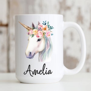 May include: White ceramic mug with a watercolor illustration of a unicorn with a floral crown. The unicorn is white with a gold horn and pink, yellow, and blue flowers in its crown. The mug has the name "Amelia" written in black cursive on the bottom.