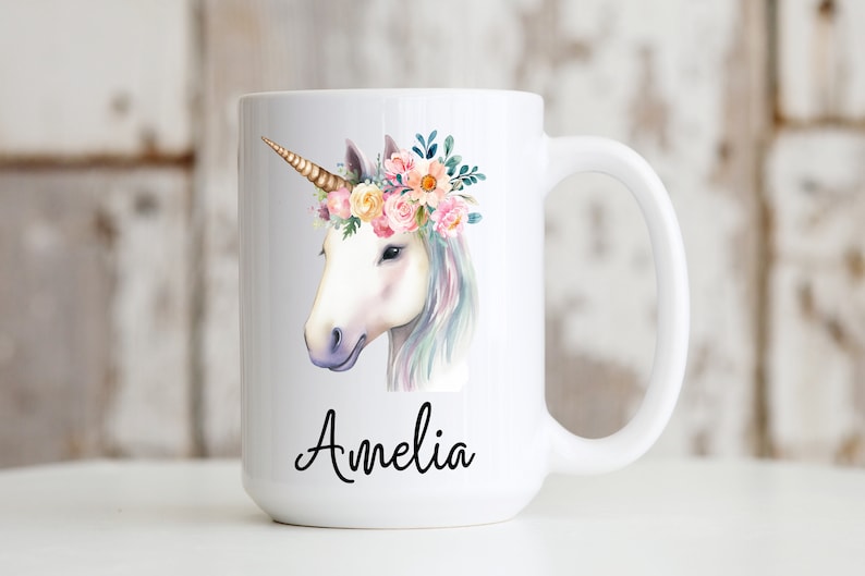May include: White ceramic mug with a watercolor illustration of a unicorn with a floral crown. The unicorn is white with a gold horn and pink, yellow, and blue flowers in its crown. The mug has the name "Amelia" written in black cursive on the bottom.