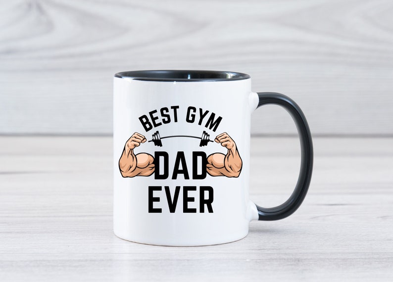 May include: White ceramic coffee mug with a black rim and handle. The mug has a black graphic that says "Best Gym Dad Ever" with a cartoon image of muscular arms lifting a barbell.