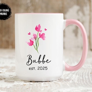 May include: White ceramic mug with a pink rim and handle. The mug features a pink floral design with the text "Bubbe est. 2025".