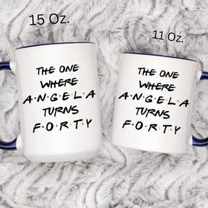 Op de afbeelding: Twee witte keramische mokken met blauwe handvatten en randen. De mokken hebben de tekst "THE ONE WHERE ANGELA TURNS FORTY" in het zwart. E&eacute;n mok is gemarkeerd met 15 Oz. en de andere met 11 Oz.