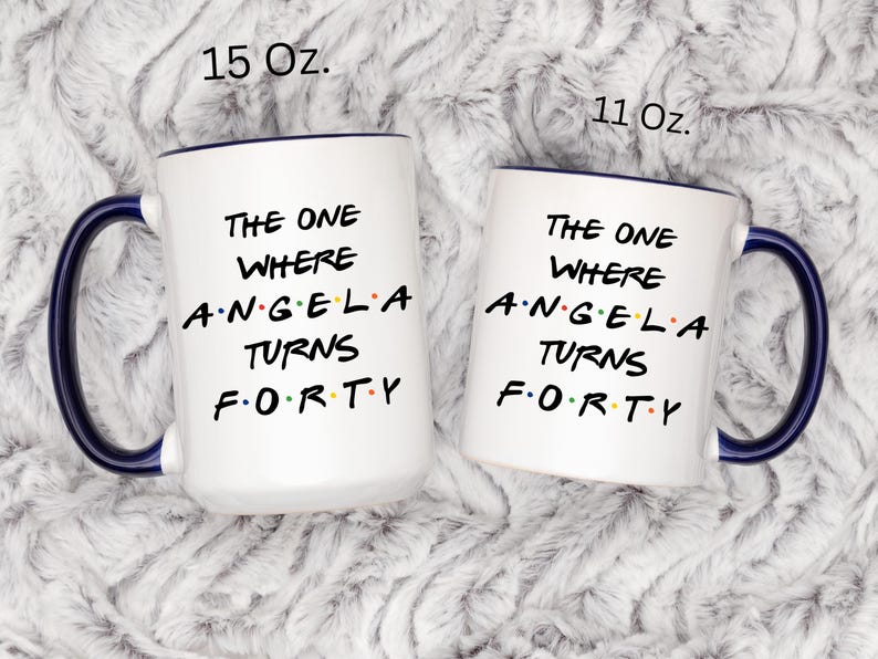 Op de afbeelding: Twee witte keramische mokken met blauwe handvatten en randen. De mokken hebben de tekst "THE ONE WHERE ANGELA TURNS FORTY" in het zwart. E&eacute;n mok is gemarkeerd met 15 Oz. en de andere met 11 Oz.