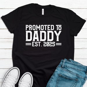 May include: Black t-shirt with white text that reads "Promoted to Daddy - Est. 2025".